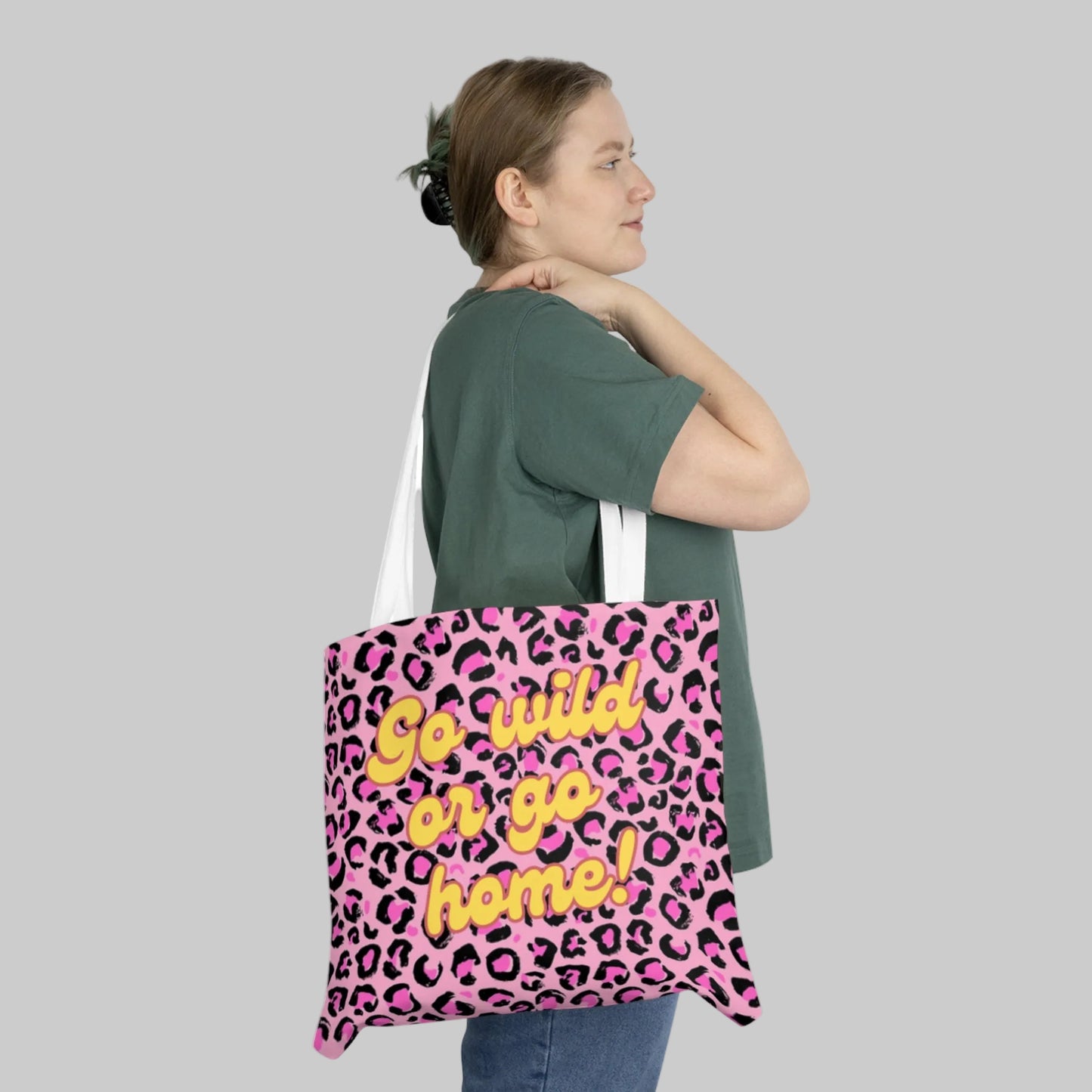Stylische Stofftasche mit auffälligem pink-schwarzem Leopardenmuster. In der Mitte steht der gelbe Schriftzug 'Go wild or go home!' in einer verspielten Schriftart. Die Tasche hat weiße Tragehenkel und wir von einer Frau im grünen T-Shirt getragen