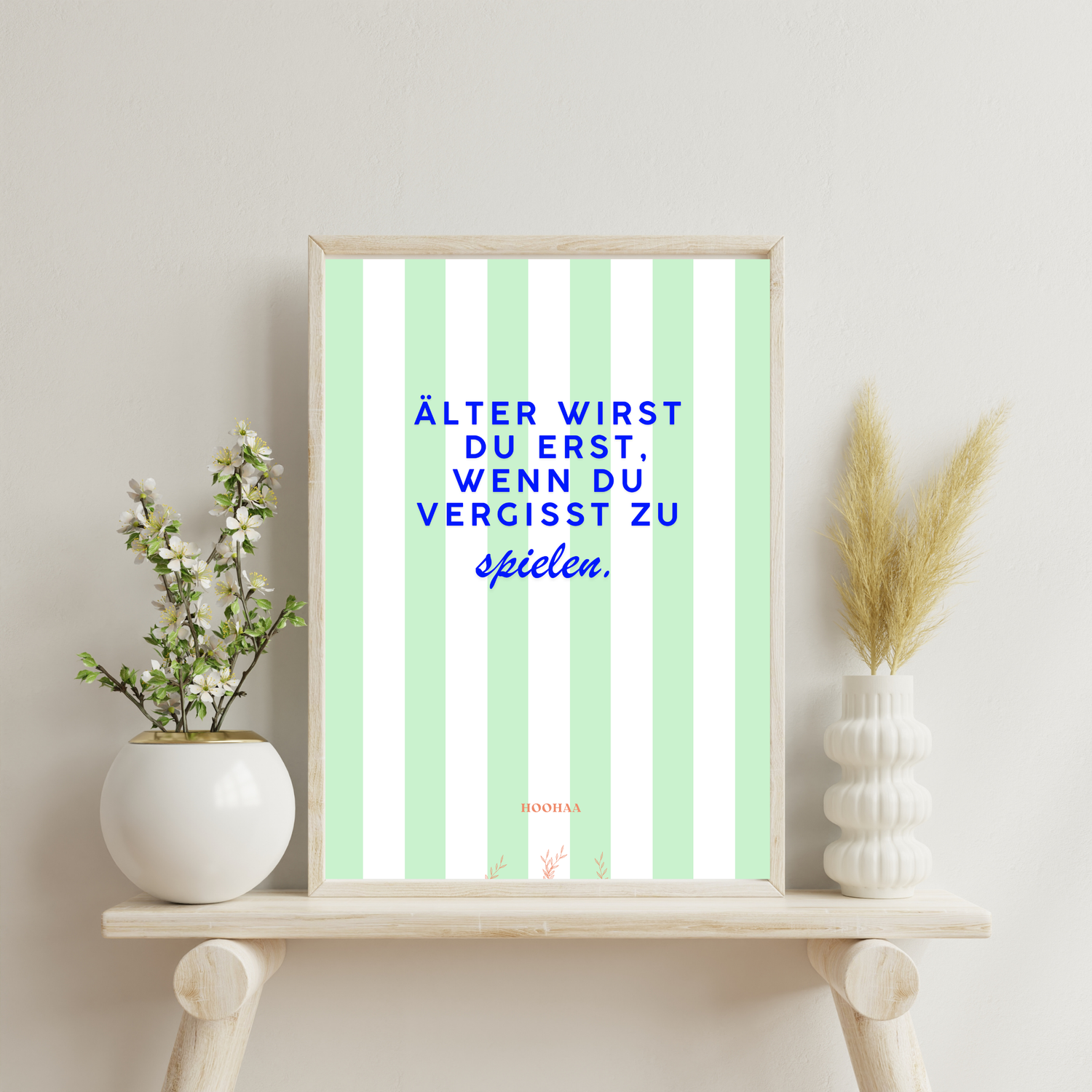 Vertikal pastellgrün und weiß gestreiftes Poster von HOOHAA, das an zwei schwarzen Klemmen und Schnüren hängt. Der Text in blauer Schrift lautet: "ÄLTER WIRST DU ERST, WENN DU VERGISST ZU" in Großbuchstaben und darunter in blauer Schreibschrift "spielen.". Am unteren Rand sind das kleine rote Logo "HOOHAA" und drei kleine rote Pflanzenzweige zu sehen. Das Poster in einem hellen Rahmen von einer Wand