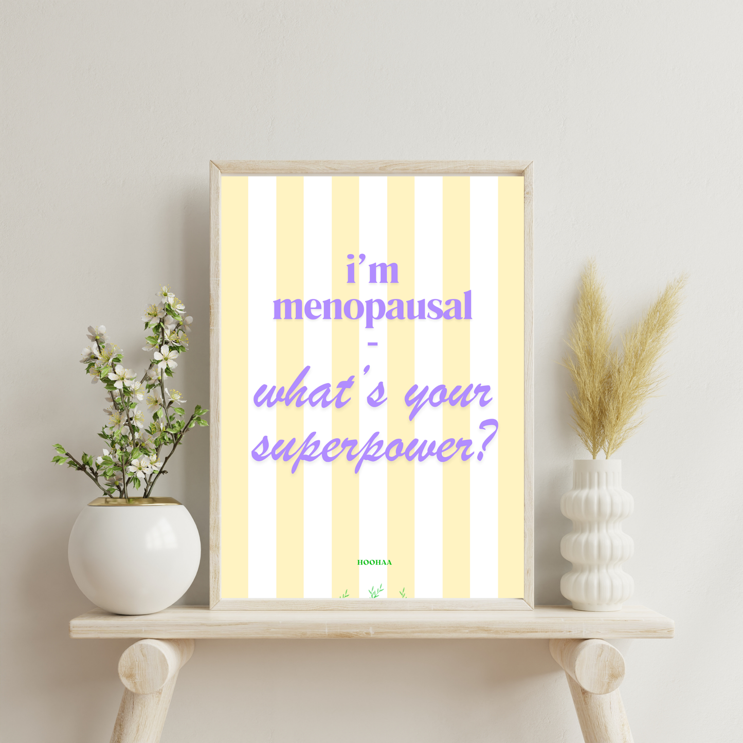 Hochformatiges Poster mit vertikalen pastellgelben und weißen Streifen. Der lila Text lautet "i'm menopausal - what's your superpower?". Am unteren Rand befindet sich der grüne Schriftzug "HOOHAA" mit kleinen pflanzlichen Illustrationen was 
auf einem weißen Sideboard an einer hellen Wand steht.
