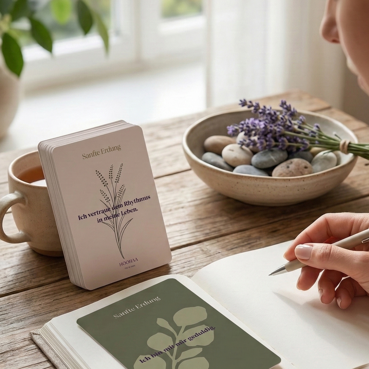 Lifestyle-Szene: Ein Stapel Affirmationskarten 'Sanfte Erdung' auf Holz und einer unterstehenden Tische Tasse Tee, Journal und Lavendel. Eine Hand-Detrakt in das Notizbuch.