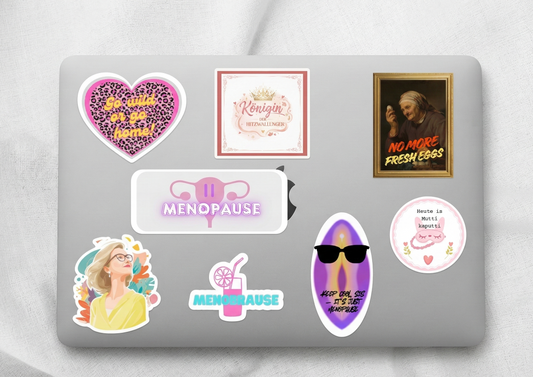 „Acht verschiedene, farbenfrohe HOOHAA Sticker mit humorvollen und wertschätzenden Motiven zur Menopause (wie ‚Königin der Hitzewallungen‘, ‚Menobrause‘ und ‚Keep cool, Sis‘), dekorativ angeordnet auf der Rückseite eines silbernen Laptops .
