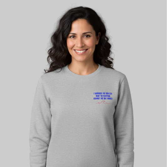 Graues Damen-Sweatshirt für die Menopause mit Rundhals ausschnitt. Auf der Rechten Brustseiten steht ein blauer Text 'I Survived the red sea now I'M FIGHTING AGAINST THE HOT WAVES' über zwei geschwungenden roten Wellen-Linien, was von einer dunkelhaarigen Frau getragen wird.