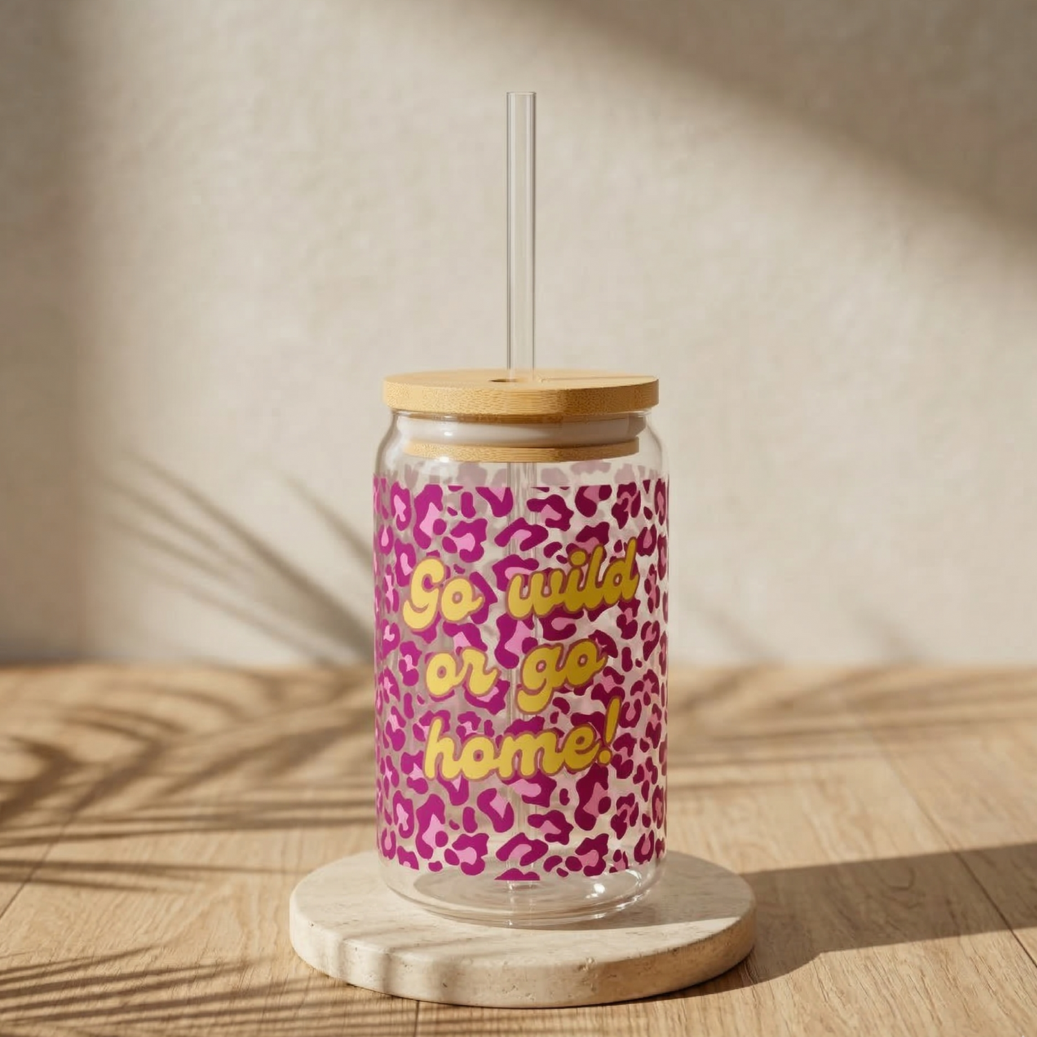 Trinkglas im Leoparden-Look in Magenta-Pink mit dem gelbenzug „Go wild or go home!“, Bambusdeckel und Glasstrohhalm auf runden Travertin-Untersätzer auf Holz mit Hintergrundblatt-Schatten im.