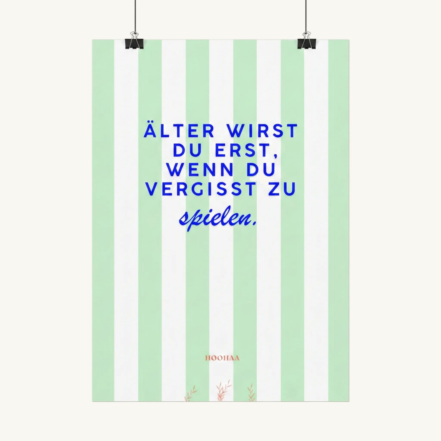 Vertikal pastellgrün und weiß gestreiftes Poster von HOOHAA, das an zwei schwarzen Klemmen und Schnüren hängt. Der Text in blauer Schrift lautet: "ÄLTER WIRST DU ERST, WENN DU VERGISST ZU" in Großbuchstaben und darunter in blauer Schreibschrift "spielen.". Am unteren Rand sind das kleine rote Logo "HOOHAA" und drei kleine rote Pflanzenzweige zu sehen.