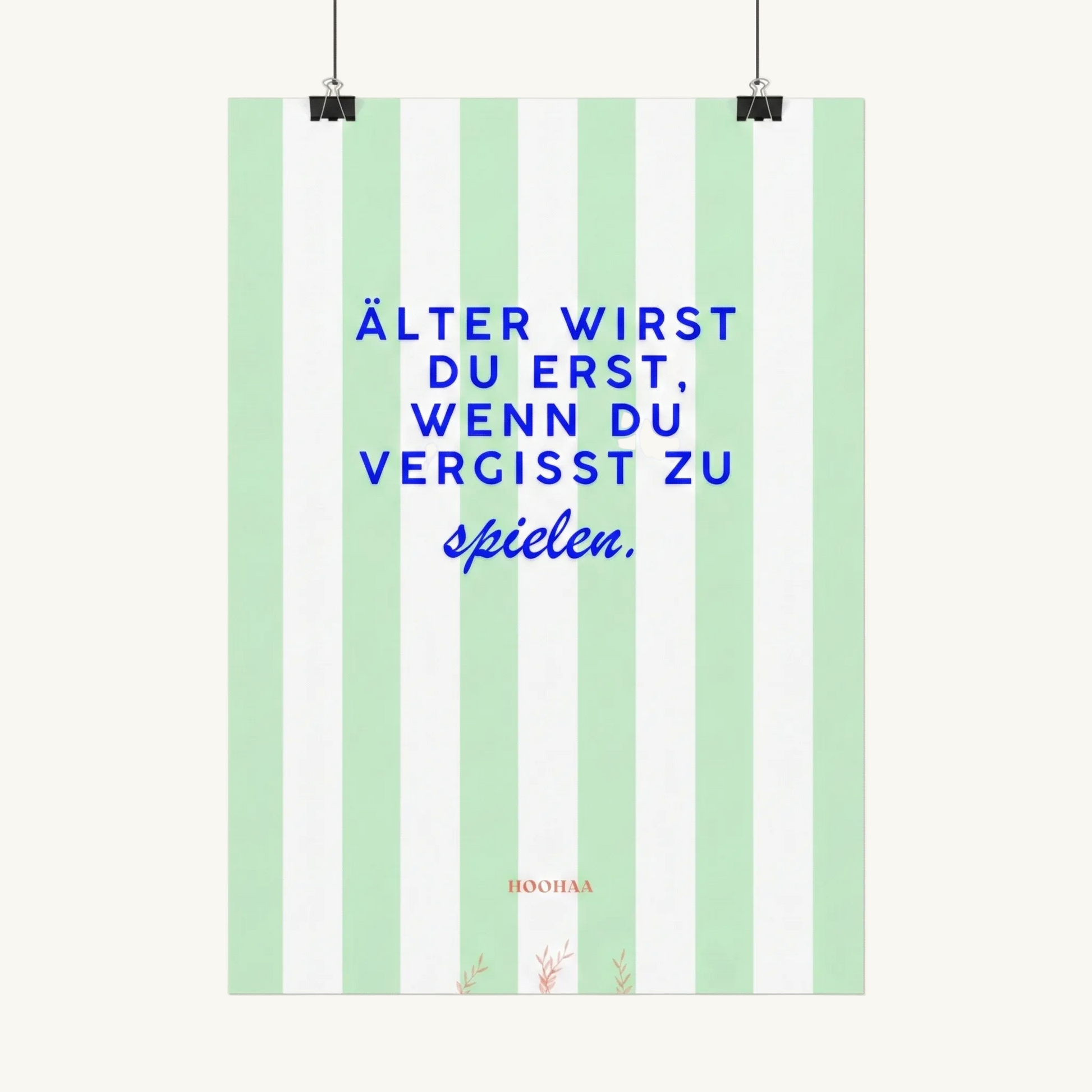 Vertikal pastellgrün und weiß gestreiftes Poster von HOOHAA, das an zwei schwarzen Klemmen und Schnüren hängt. Der Text in blauer Schrift lautet: "ÄLTER WIRST DU ERST, WENN DU VERGISST ZU" in Großbuchstaben und darunter in blauer Schreibschrift "spielen.". Am unteren Rand sind das kleine rote Logo "HOOHAA" und drei kleine rote Pflanzenzweige zu sehen.