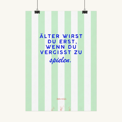 Vertikal pastellgrün und weiß gestreiftes Poster von HOOHAA, das an zwei schwarzen Klemmen und Schnüren hängt. Der Text in blauer Schrift lautet: "ÄLTER WIRST DU ERST, WENN DU VERGISST ZU" in Großbuchstaben und darunter in blauer Schreibschrift "spielen.". Am unteren Rand sind das kleine rote Logo "HOOHAA" und drei kleine rote Pflanzenzweige zu sehen.