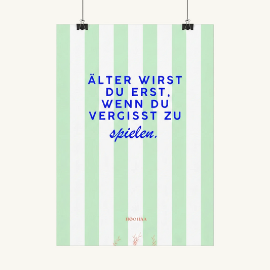 Vertikal pastellgrün und weiß gestreiftes Poster von HOOHAA, das an zwei schwarzen Klemmen und Schnüren hängt. Der Text in blauer Schrift lautet: "ÄLTER WIRST DU ERST, WENN DU VERGISST ZU" in Großbuchstaben und darunter in blauer Schreibschrift "spielen.". Am unteren Rand sind das kleine rote Logo "HOOHAA" und drei kleine rote Pflanzenzweige zu sehen.