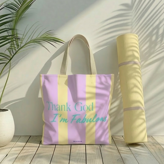  Stofftasche mit lila-gelben Streifen und dem tSpruch "Thank God I'm Fabulous" von HOOHAA, die neben einer gelben Yogamatte vor einer hellen Zimmerwand steht.