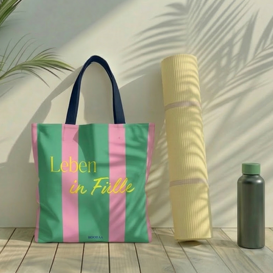 Tote Bag mit grün-pinken Streifen und dem gelben Schriftzug „Leben in Fülle“ von HOOHAA, im Setting mit einer Palme, heller Yogamatte und Trinkflasche bei ästhetischem Lichteinfall.