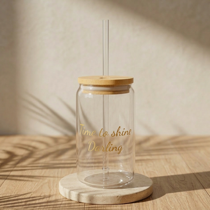 Ein minimalistisches, transparentes Glas in Dosenform mit hellen Holzdeckel und einem Glasstrohhalm. Auf der Vorderseite ist ein Spruch in goldener Schreibschrift „Time to shine Darling“. Das Glas steht vor einer schlichten, weißen Hintergrund.