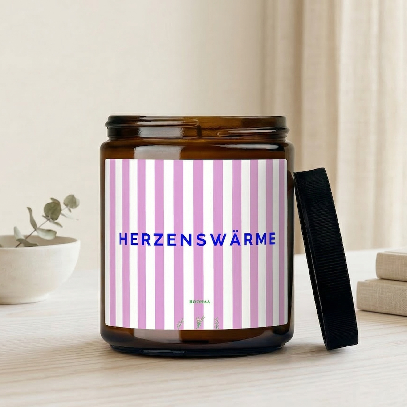 Handgefertigte Sojawachskerze im Braunglas mit rosa-weiß gestreiftem Etikett und dem Schriftzug „Herzenswärme“, platziert auf einem hellen Holztisch neben einem Topf Lavendel in einem freundlich beleuchteten Wohnzimmer.