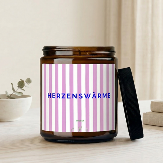  Handgefertigte Sojawachskerze im Braunglas mit rosa-weiß gestreiftem Etikett und dem Schriftzug „Herzenswärme“, platziert auf einem hellen Holztisch neben einem Topf Lavendel in einem freundlich beleuchteten Wohnzimmer.