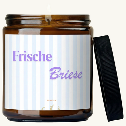 Hochwertige handgefertigte Sojawachs-Duftkerze „Frische Briese“ in einem Braunglas, hergestellt in Berlin, mit blau-weiß gestreiftem Etikett vor einer hellen Wand.
