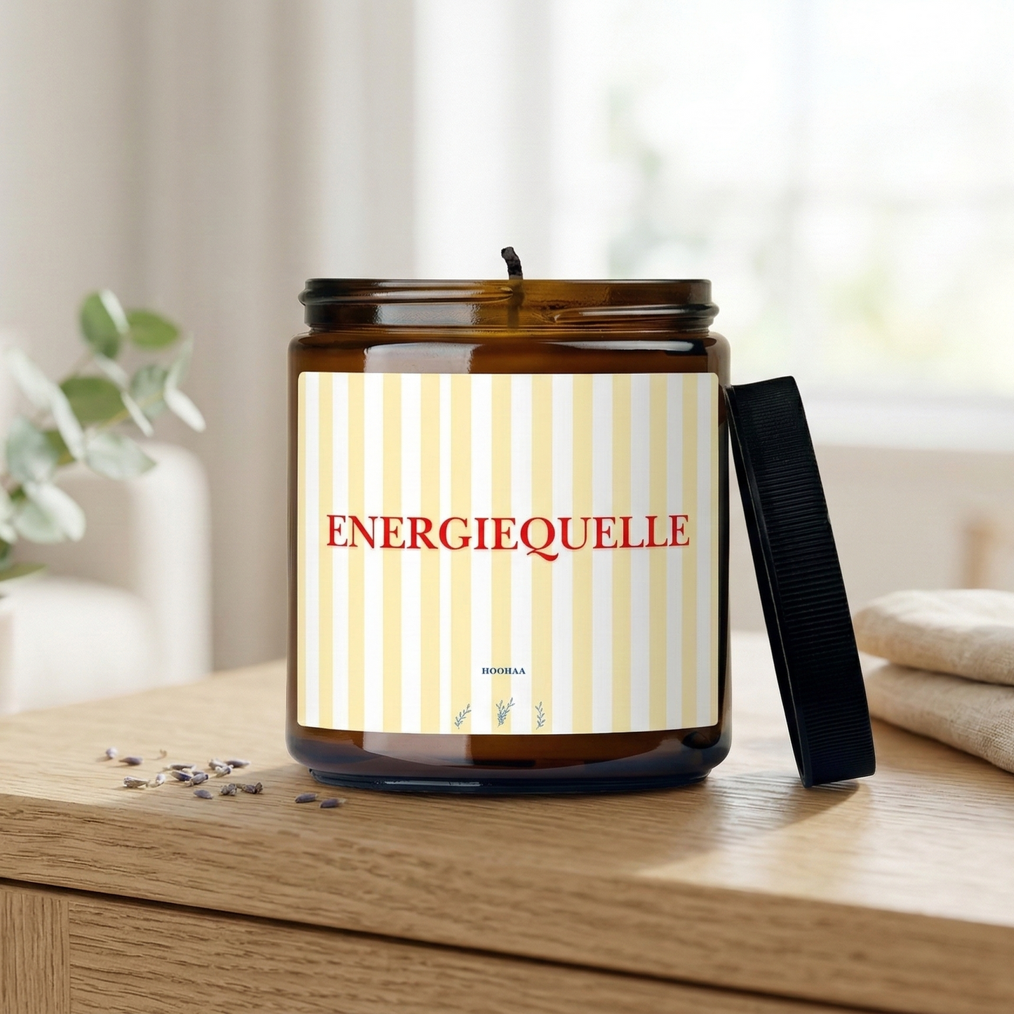 Mockup einer handgefertigten Sojawachskerze im Braunglas mit gelb-weiß gestreiftem Etikett und dem roten Schriftzug „Energiequelle“, platziert in einer hellen, modernen Wohnumgebung auf einem Eichentisch.