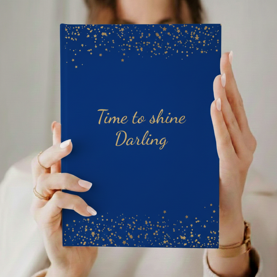 Blaues A5 Notizbuch mit der Aufschrift 'Time to shine Darling' in goldener Schrift mit kleinen goldenen Sternen für die Menopause. Das Journal ist als Begleiter für Achtsamkeit und Reflexion für Frauen in den Wechseljahren konzipiert was von einer Frau direkt in die Kameragehalten wird.