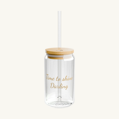 Ein minimalistisches, transparentes Glas in Dosenform mit hellen Holzdeckel und einem Glasstrohhalm. Auf der Vorderseite ist ein Spruch in goldener Schreibschrift „Time to shine Darling“. Das Glas steht vor einer schlichten, weißen Hintergrund.
